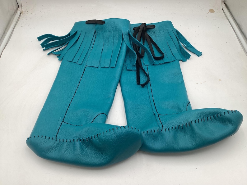 May include: A pair of turquoise leather moccasin boots with fringe detail at the top. The boots have a knee-high design with black ties and stitching. The soles are stitched with a contrasting thread.