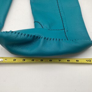 May include: Turquoise leather moccasin-style baby booties with black stitching. The booties are approximately 10 inches long, as measured by a yellow tape measure. The leather has a textured appearance.