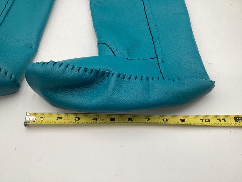 May include: Turquoise leather moccasin-style baby booties with black stitching. The booties are approximately 10 inches long, as measured by a yellow tape measure. The leather has a textured appearance.