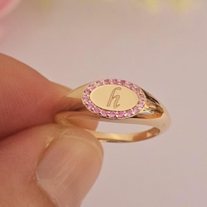 Op de afbeelding: Een goudkleurige zegelring met een ovaalvormig vlak waarin in het midden een kleine letter h is gegraveerd. Het ovale vlak is omringd door een rand van kleine, ronde, roze steentjes die in het metaal zijn gezet als een decoratief kader.