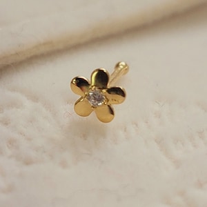 May include: Gold nose ring with a small flower design and a clear gemstone in the center.