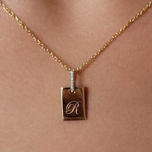 Könnte beinhalten: Eine goldene Kettenkette mit einem kleinen rechteckigen Anhänger, der mit einem diamantbesetzten Rand und dem eingravierten Buchstaben "R" in der Mitte verziert ist.