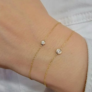 May include: Two delicate gold chain bracelets, each featuring a single round, bezel-set clear gemstone. The thin, polished gold chains are worn together, showcasing the minimalist design of the circular stone settings against the skin.