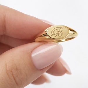 May include: A gold signet ring featuring an oval face engraved with a cursive letter B. The ring has a polished, smooth finish and a tapered band. The design is simple and classic, showcasing the metallic luster of the gold material throughout.