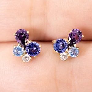 Könnte beinhalten: Ein Paar goldene Ohrstecker mit einer Ansammlung verschiedenfarbiger Edelsteine. Jeder Ohrring besteht aus einem birnenförmigen violetten Stein, einem runden dunkelblauen Stein, einem runden hellblauen Stein und einem kleinen klaren weißen Stein.