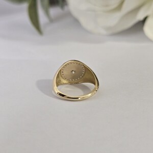 May include: A gold signet ring with a circular face. The face features a central diamond encircled by a halo of smaller diamonds. The ring is set against a soft, neutral background.