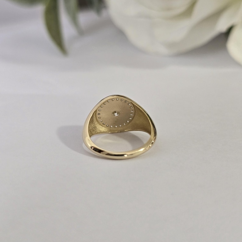 May include: A gold signet ring with a circular face. The face features a central diamond encircled by a halo of smaller diamonds. The ring is set against a soft, neutral background.