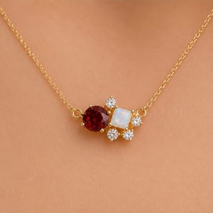 May include: A gold chain necklace featuring a cluster pendant with a round deep red garnet, a square white opal, and five small clear round stones arranged in a floral pattern. The metal setting is gold-toned and holds all the gemstones securely.