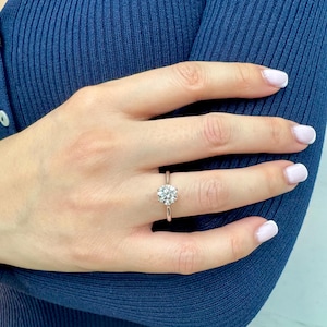 May include: A silver band engagement ring featuring a round-cut diamond. The ring is displayed on a hand with manicured nails, set against a dark blue ribbed fabric. The diamond is the focal point.