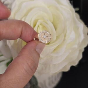 May include: A gold signet ring with a starburst design, a central diamond, and a halo of smaller diamonds. The ring has a polished finish. The ring is held against a backdrop of a white rose.