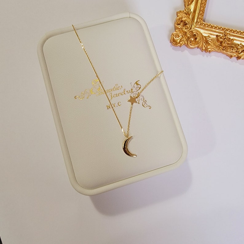 May include: A gold crescent moon pendant necklace with a small star charm. The necklace is in a white box with the words "Jewellery" and "NYC" printed on it.