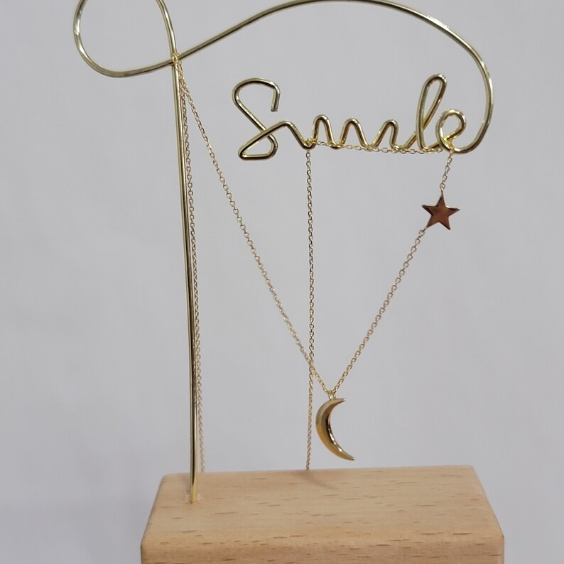 May include: Gold metal necklace display stand with the word "Smile" written in cursive. The stand has two chains with a crescent moon and a star pendant.