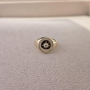 May include: A gold ring with a starburst design. The ring features a central star-shaped stone surrounded by a circle of smaller stones. The band is a solid gold colour, and the ring is set against a neutral background.