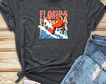 Puede incluir: Una camiseta gris oscuro con un gráfico de una persona surfeando en una ola. El gráfico está en un estilo retro con la palabra "FLORIDA" repetida en diferentes colores.