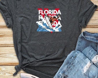 May include: A dark gray t-shirt with a graphic of a surfer riding a wave with the word "Florida" repeated three times in red and white letters.