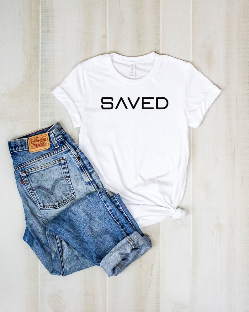 May include: White t-shirt with the word "SAVED" printed in black on the front. The shirt is laying on a light wood surface with a pair of blue jeans.