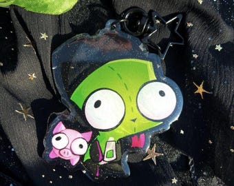 May include: A green cartoon character with large eyes and a pink pig character on a black background with gold glitter stars. The characters are on a clear acrylic keychain with a black metal ring.