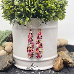 May include: A pair of teardrop-shaped dangle earrings featuring a multi-colored confetti pattern in shades of pink, orange, white, and black. The earrings are suspended from silver-toned metal hooks and have a smooth, glossy finish on the surface.