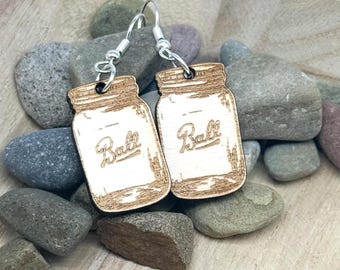 May include: A pair of wooden earrings shaped like mason jars with the word "Ball" engraved on them. The earrings have silver-colored hooks and are displayed on a wooden tray with small stones.