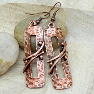 May include: A pair of copper-toned earrings featuring a rectangular frame with a hammered texture. Each earring has a central open space crossed by two thin, curved metal bars accented with small spherical beads and attached to a hook wire.