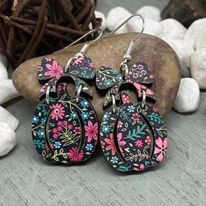 May include: A pair of wooden pumpkin earrings with a floral pattern in pink, blue, and green on a black background. Each earring has a bow-shaped top and a pumpkin-shaped bottom, connected by a silver hook.