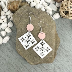 May include: A pair of earrings with a floral design. Each earring features a small, light pink circle connected to a white, diamond-shaped pendant with a cut-out floral pattern. The earrings have silver-colored hooks.