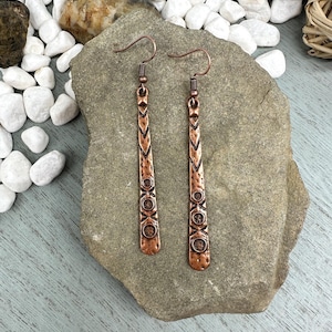 May include: A pair of copper-colored earrings with a linear design. The earrings have a hook closure and a series of geometric shapes and patterns. The earrings are displayed on a gray stone surface.