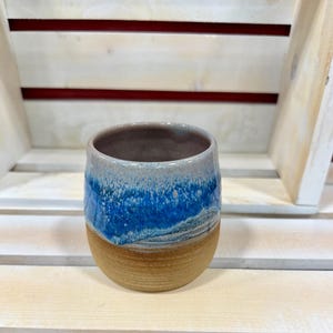 May include: A handmade ceramic tumbler with a unique design. The top half features a vibrant blue glaze that transitions to a lighter, speckled blue, while the bottom half is a warm, earthy brown. The interior is a muted gray.