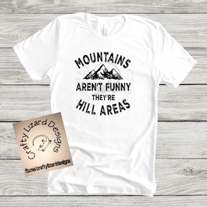 Puede incluir: Camiseta blanca con un gr&aacute;fico negro que dice "Mountains aren't funny they're hill areas" con una silueta de monta&ntilde;as.