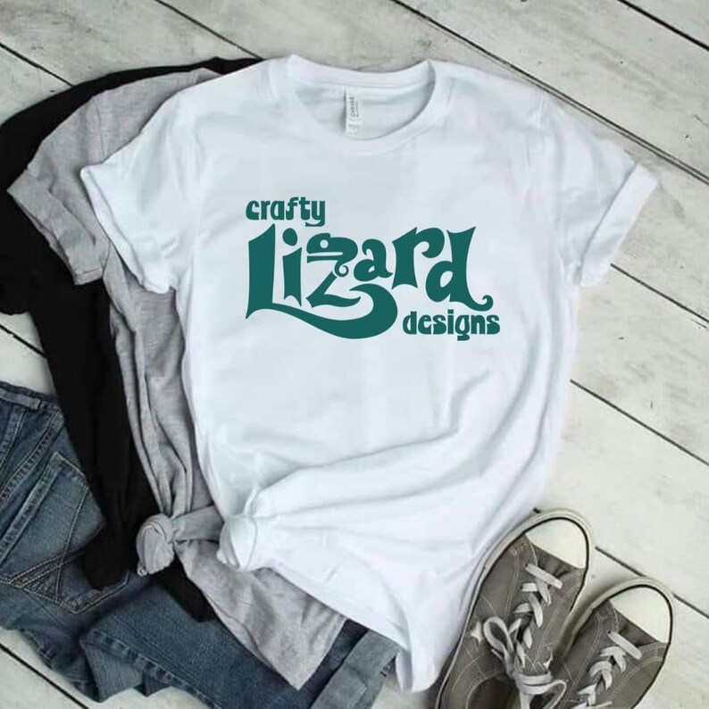 Puede incluir: Camiseta blanca con un logotipo verde azulado que dice "crafty lizard designs".
