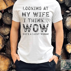 May include: White t-shirt with the text "LOOKING AT MY WIFE I THINK... WOW SHE'S A LUCKY WOMAN" in a black, distressed font. The shirt is short-sleeved and worn by a person with their hands in their pockets.