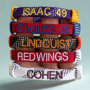 May include: A stack of personalized bracelets in various colors. The bracelets are embroidered with names and numbers, including "ISAAC 49", "EAGLES 25", "LINDQUIST", "RED WINGS", and "COHEN". The bracelets are made of a woven material.