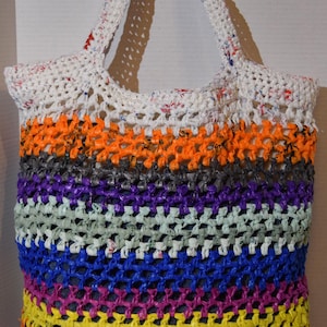 May include: A colorful crocheted tote bag made from recycled plastic. The bag has a white base with stripes of orange, gray, purple, blue, yellow, green, and red.