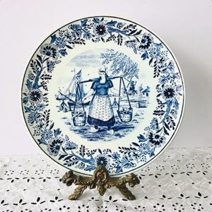 May include: A white and blue decorative plate featuring a Dutch woman carrying buckets, surrounded by a floral border. The plate has a diameter of approximately 10 inches. The design includes windmills and sailboats.