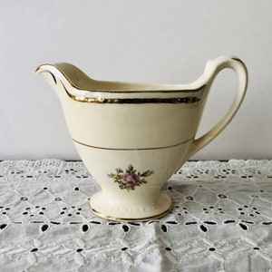 May include: A white ceramic creamer with a gold trim and a floral design. The creamer has a small, round base and a handle.