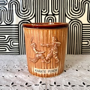 May include: A brown ceramic tumbler with a textured, vertical-line design. The tumbler features embossed figures in karate poses and the text "KARATE PUNCH" on a white rectangular label. The rim is a darker brown.