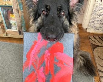 May include: A German Shepherd dog holds a canvas painting with abstract red and pink paint strokes. The painting is titled "MODERN LOVE".