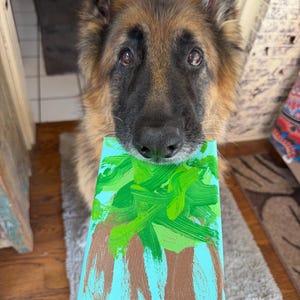 Pode incluir: Um cão pastor alemão de pelo comprido segurando uma tela retangular na boca. A tela apresenta uma cena pintada à mão com um fundo azul claro, troncos de árvore castanhos e pinceladas de verde vivo representando a copa da árvore.