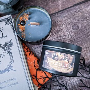 May include: Two black candles with labels. The candle on the left has a black wax with orange and brown accents. The candle on the right has a label that reads "Black Grimoire" with the text "Soy & Essential Oil Candle" underneath.