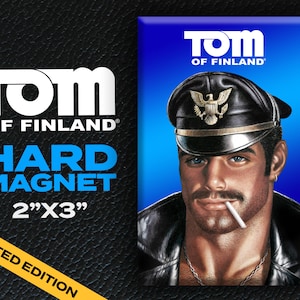 May include: A blue and white hard magnet featuring a man wearing a black leather cap with a gold eagle emblem, a black leather jacket, and a cigarette in his mouth. The magnet is 2 inches by 3 inches and says "Tom of Finland" in white letters on a blue background.