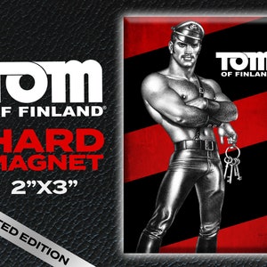 Op de afbeelding: Een zwart-witte illustratie van een man in een leren outfit en een pet. Hij houdt een sleutelbos vast. De afbeelding is op een rood-zwarte gestreepte achtergrond met de tekst "TOM OF FINLAND" in wit. De tekst "HARD MAGNET 2"X3" LIMITED EDITION" is ook zichtbaar.