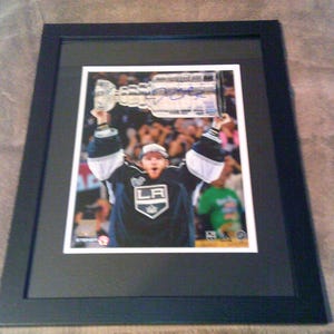 May include: Framed photograph of a hockey player holding a trophy above his head. The player is wearing a black and white jersey with the letters "LA" on the front. The photograph is in a black frame.