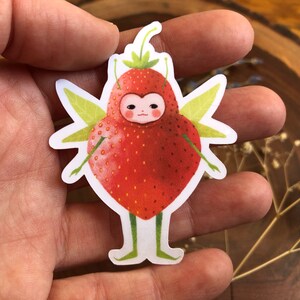 Pu&ograve; includere: Una fragola rossa con un viso da cartone animato carino e ali verdi. La fragola indossa un vestito bianco e ha delle piccole gambe bianche.