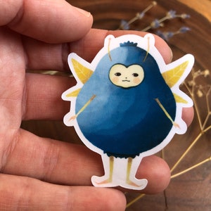 Pu&ograve; includere: Un adesivo di cartone animato blu e giallo con un mirtillo con le ali e una faccia. Il mirtillo indossa un costume blu e giallo.