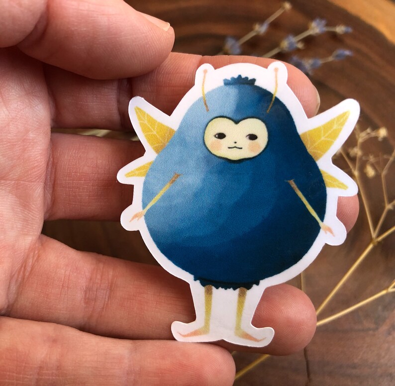 Pu&ograve; includere: Un adesivo di cartone animato blu e giallo con un mirtillo con le ali e una faccia. Il mirtillo indossa un costume blu e giallo.