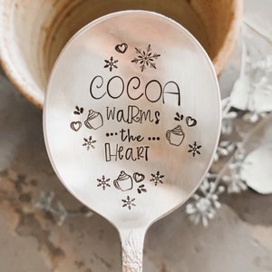 May include: A silver spoon with the words "COCOA warms the Heart" etched on the bowl, decorated with snowflakes and hearts. The spoon is near a ceramic mug, evoking a cosy, winter theme.