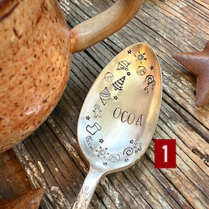 May include: A silver spoon with the word "OCOA" and Christmas-themed illustrations, including a Christmas tree and gingerbread men. A brown mug with a handle is in the background, along with a red ribbon and a star.
