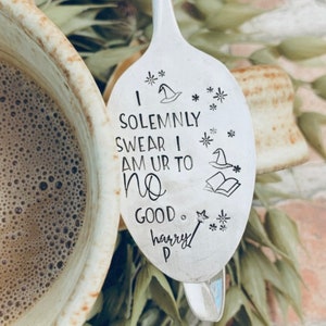 May include: A silver spoon with the text "I SOLEMNLY SWEAR I AM UR TO NO GOOD. harry P" and illustrations of a wizard hat, stars, a book, and a wand. The spoon is surrounded by greenery and a cup of coffee.
