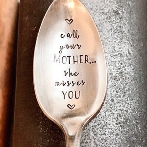 May include: A silver spoon with the words "call your MOTHER... she misses YOU" etched on the bowl. The text is in a handwritten style, with a heart symbol above and below the text. The spoon is set against a textured background.