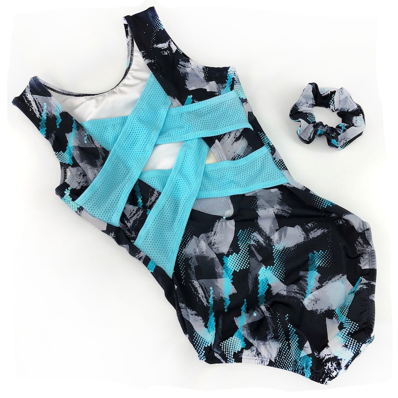 May include: A black leotard with a blue and grey abstract print. The leotard has a criss-cross back design with a blue mesh panel. A blue hair scrunchie is included.